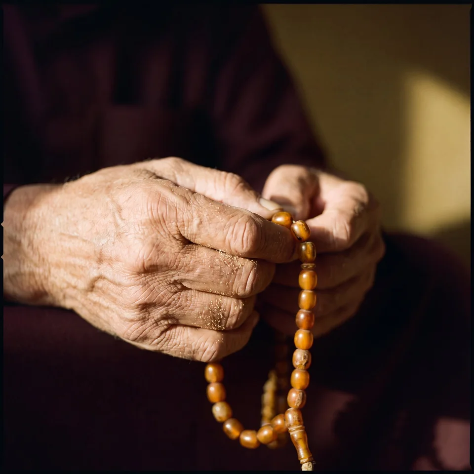 A close view of weathered hands holding amber-coloured prayer beads against dark red fabric — generative artwork.