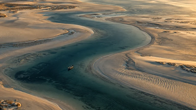 An overhead view of a winding tidal channel, pale sandbars and a small wooden boat — AI-generated artwork.